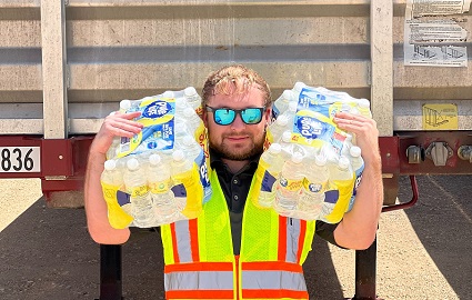 Man in mirrored sunglasses holding two cases of bottled water perched on each shoulder, standing in front of XTRA Lease flatbed trailer